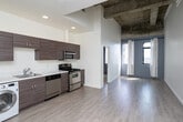 Located at 3075 Wilshire Blvd in the vibrant Koreatown neighborhood of Los Angeles, this unit blends industrial character with modern updates. The living area makes a strong architectural statement with soaring ceilings that reveal exposed concrete beams, paired with light hardwood flooring and crisp white walls that create a bright, open atmosphere. Large windows frame dynamic views of the surrounding Los Angeles cityscape, filling the space with abundant natural light throughout the day. The bedroom features soft carpeting, additional city-facing windows with sheer curtains, and the same airy, well-lit quality found throughout the unit. The kitchen is thoughtfully designed with white cabinetry, light countertops, and a full suite of stainless steel appliances including a refrigerator, dishwasher, and range. A washer unit is conveniently integrated into the kitchen layout, adding practical in-unit laundry functionality. A full bathroom completes the floor plan. The two-toned gray and white kitchen walls add a contemporary design touch that complements the overall modern-industrial aesthetic of the space. Situated along one of Los Angeles' most recognized corridors, this unit offers easy access to the dining, retail, and transit options that define the Koreatown and Mid-Wilshire area.
