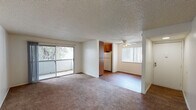 This single-floor unit offers a practical and well-organized layout designed for comfortable everyday living. The open-plan living and dining area is generously proportioned, featuring wall-to-wall carpeting, a textured ceiling, and a ceiling fan that enhances airflow throughout the space. A sliding glass door leads directly to a private balcony, bringing in natural light and offering pleasant views of the surrounding greenery. The living area flows seamlessly into the dining space, which connects to the galley-style kitchen. The kitchen is equipped with warm wood-toned cabinetry, a white refrigerator, a range, a dishwasher, and a sink, all set against durable hardwood-style flooring. Two bedrooms are each carpeted and bathed in natural light through large windows that frame outdoor tree views, creating a calm and restful atmosphere. The bathroom features a wide vanity with an oversized mirror, ample counter space, and a separate toilet and shower/tub combination. Clean white walls and neutral tones throughout the unit contribute to a bright, cohesive aesthetic that is both functional and inviting.