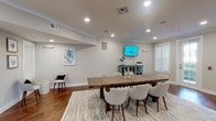 This single-floor space is thoughtfully designed with a coastal-inspired aesthetic, combining functionality with a polished, welcoming atmosphere. The interior is finished throughout with rich hardwood flooring that anchors the neutral, soft-gray palette of the walls and crisp white trim, creating a cohesive and professional environment.

The central gathering area serves as the focal point of the space, featuring a large rustic wood farmhouse table surrounded by upholstered white chairs set atop a textured area rug. A wall-mounted flat-screen display and a distressed blue sideboard add both utility and character to the room. Recessed lighting illuminates the space evenly, complemented by natural light filtering through white-slatted window blinds.

A cozy seating nook off the main area provides an intimate setting for smaller conversations, furnished with two upholstered chairs, a round textured side table, and coastal wall art depicting coral in muted blues and greens.

Additional areas of the space include private rooms accessible through glass-paneled French doors with integrated blinds, offering privacy while maintaining visual openness. A service or reception window is built into one wall, suggesting a client-facing function. Crown molding throughout adds an elevated architectural detail, reinforcing the space's refined yet approachable character.