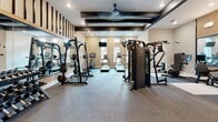 This fitness center offers a thoughtfully designed single-floor layout that accommodates a comprehensive range of workout activities. The space is organized into distinct functional zones, maximizing utility while maintaining an open, uncluttered feel. Neutral-toned walls, recessed lighting, and dark structural beam accents create a clean, modern aesthetic throughout.

The main training area features a variety of strength equipment, including cable machines, a multi-functional power rack, adjustable benches, and a full dumbbell rack. Large mirrors line the walls, enhancing the sense of space and allowing users to monitor their form. A dedicated cardio section houses multiple treadmills and an elliptical trainer, positioned near windows that bring in natural light. A ceiling fan overhead provides airflow and comfort during intense workouts.

A separate functional training zone is defined by a boxing-focused setup, complete with heavy punching bags suspended from a sturdy overhead rig, a plyo box, medicine balls, a BOSU ball, and additional free weights. This area benefits from vaulted ceiling height, adding an airy, open quality.

Durable, low-pile carpet tiles cover the floors throughout, offering traction and comfort. Glass-paneled doors connect to an adjacent room, and the overall ambiance is professional, clean, and purpose-driven.