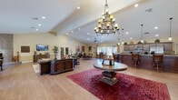 This single-story community clubhouse is designed to offer a welcoming and versatile gathering space for residents. The heart of the facility is a sweeping open-plan great room with soaring vaulted ceilings, rich hardwood-style flooring, and an elegant chandelier as a centerpiece. Recessed lighting and ceiling fans complement the natural light streaming through multiple windows dressed with decorative curtains. A welcoming entryway with glass double doors and a branded entry mat sets the tone upon arrival, flanked by comfortable leather seating areas with tall indoor plants adding a touch of greenery.

The full kitchen is outfitted with dark wood cabinetry, granite countertops, stainless steel appliances, and a large island with bar seating—ideal for hosting events or casual gatherings. A separate dining area features a striking floor-to-ceiling mosaic tile fireplace that serves as a dramatic focal point, accompanied by ample seating and natural views through large windows.

A game room provides additional recreational space, complete with a pool table and a casual seating arrangement. The dedicated fitness center is equipped with cardio machines, a cable strength system, free weights, and wall mirrors, all set on durable rubber-tiled flooring with natural light from surrounding windows.