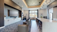 Located at 100 Forge Circle in Lawrence Township, NJ, this thoughtfully designed communal kitchen and living room offers a refined, upscale atmosphere ideal for gathering and entertaining. The space showcases a striking tray ceiling with integrated ambient lighting and geometric pendant fixtures that serve as a focal point throughout the room.

The kitchen is appointed with sleek white cabinetry, a lengthy quartz-topped island with bar seating, stainless steel appliances including a full-size refrigerator, and a stylish tile backsplash. Geometric-patterned floor tiles extend through the kitchen area, adding visual interest and a contemporary touch.

The living area transitions seamlessly into a comfortable lounge featuring plush leather-style armchairs arranged to face a large wall-mounted television set within a dark wood accent surround, complemented by white lower cabinetry. Patterned carpeting defines this seating zone, adding warmth and texture.

A mix of intimate seating options—including curved booth-style chairs with marble-top tables and bar-height counter seating—offer versatile arrangements suited to both casual conversation and work. Large windows dressed with draping curtains flood the space with natural light and provide views of the outdoors. Curated abstract artwork accents the walls, enhancing the modern and inviting ambiance throughout.