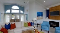 Located at 500 Talon Dr in Jacksonville, NC, this single-story residence makes a strong architectural statement with its soaring vaulted ceilings and striking arched transom window that floods the main living area with natural light. The living room serves as the centerpiece of the home, showcasing the impressive ceiling height and an open, airy layout that creates a sense of generous space. Large French doors provide a seamless connection to the outdoors, offering views of what appears to be a pool area, further enhancing the bright and welcoming atmosphere. The interior design carries a vibrant coastal character, with crisp white walls accented by bold pops of color through furnishings and artwork, including vivid blues, greens, and reds that enliven the space. Recessed lighting complements the abundant natural light throughout. Crown molding and clean architectural lines add a refined, polished finish to the home's interior. The layout is thoughtfully arranged on a single level, offering comfortable and accessible flow between living spaces. Overall, this home combines architectural elegance with a cheerful, light-filled ambiance well-suited to the coastal Carolina lifestyle.