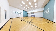 This indoor sports facility centers on a generously sized hardwood court finished in warm maple tones, with crisp multi-sport line markings laid out for basketball, volleyball, and pickleball. The court is equipped with two regulation basketball hoops mounted on ceiling-suspended structures, providing unobstructed play space below. Padding along the walls near each baseline ensures player safety during competitive use.

The ceiling soars overhead, creating an open, airy atmosphere enhanced by rows of flush-mounted fluorescent light panels that provide consistent, even illumination across the entire court surface. A series of windows along one wall allows natural light to filter into the space, further brightening the interior.

Bold "Home Team" and "Visitor Team" signage is painted directly onto teal and white cinder block accent walls at each end of the court, adding a spirited, competitive character to the design. A large glass window on one side of the court looks directly into an adjacent fitness room equipped with cardio machines and strength training equipment, creating a visual connection between the two athletic spaces. The overall design is clean, functional, and purpose-built for a wide range of indoor recreational and competitive activities.