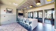 Located at 4820 SW 48th Ave in Ocala, Florida, this single-floor professional workspace offers a thoughtfully designed environment ideal for productivity and collaboration. The space is anchored by a spacious conference room featuring a long white marble-topped table surrounded by modern ergonomic chairs, a wall-mounted flat-screen display, and distinctive drum pendant lighting with gold interiors. A striking wood-paneled ceiling with a chevron pattern adds warmth and architectural character to the conference area, complemented by two-tone sage green and neutral wall treatments and classic wainscoting. Flanking the main area are two private office suites enclosed by full-height black-framed glass doors, providing visual transparency while maintaining privacy. Each private office is furnished with a desk and seating, and benefits from large windows that frame views of the surrounding Florida landscape, including swaying palm trees. A corridor lined with expansive windows floods the interior with natural light, leading to a business support station equipped with a printer and cabinetry. Throughout the space, geometric patterned flooring — a dynamic mix of wood-look tile laid in a herringbone mosaic — creates a visually striking foundation that ties the entire layout together. The overall ambiance is modern, polished, and professional.