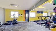 This fitness center is a thoughtfully designed, single-level workout facility that combines energetic aesthetics with a comprehensive range of exercise equipment. The space is immediately distinguished by its bold color palette — vivid yellow walls accented by striking orange and navy blue vertical stripes — creating a dynamic and motivating atmosphere throughout.

The facility is organized into clearly defined workout zones. The main cardio area houses multiple Echelon treadmills and stationary cycling bikes, positioned beneath wall-mounted flat-screen televisions and complemented by full-length mirrors that visually expand the space. A dedicated strength training section features a multi-tier dumbbell rack with a full range of free weights and an adjustable workout bench set on protective floor matting. An elliptical trainer and additional cardio equipment round out the machine offerings.

A separate area provides space for functional training, complete with a rack of medicine balls and a Reflect smart mirror for guided workouts. Natural light filters in through multiple windows, offering views of the surrounding outdoor area. The textured carpet tile flooring runs throughout, while flush-mount and circular ceiling light fixtures provide consistent, even illumination. The space is clean, well-maintained, and equipped with a fire safety station, ensuring a safe and functional environment for all users.