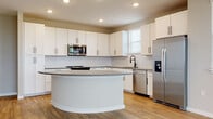 Located on Keymar Drive in Davenport, Florida, this single-story home presents a clean, contemporary design with a neutral color palette and consistent finishes throughout. The kitchen serves as a central highlight, featuring a distinctive curved island with gray countertops, white shaker-style cabinetry, a white subway tile backsplash, and a full suite of stainless steel appliances including a refrigerator with ice dispenser, built-in microwave, range, and dishwasher. Natural light enters through multiple windows fitted with horizontal blinds, complementing the warm wood-look luxury vinyl plank flooring that extends through the main living and kitchen areas. The open layout connects the kitchen to a spacious living area, providing a generous, functional flow. Bedrooms are finished with plush neutral carpet and offer walk-in closet access. The primary bathroom is well-appointed with a double-sink vanity featuring gray countertops, white cabinetry, matte black fixtures, framed mirrors, and a glass-enclosed shower. A secondary bathroom offers a tiled tub-shower combination with matte black hardware. An additional versatile room with wood-look flooring and a window provides flexible use as a home office or utility space.