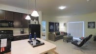 This single-floor residence offers a thoughtfully designed open-concept layout that creates a seamless flow between the kitchen, dining, and living spaces. The kitchen is outfitted with dark espresso cabinetry, black appliances including a full-size refrigerator, and a light-toned countertop that doubles as a breakfast bar. Elegant pendant lighting hangs above the counter, adding a stylish focal point to the space. The open living area features plush carpeting and is furnished with a curved sofa and ottoman, anchored by a pair of bold framed art prints that lend a contemporary aesthetic. Vertical blinds along the far wall allow for flexible natural light control. A glimpse into the adjacent bedroom reveals a cozy, carpeted retreat with soft furnishings, maintaining the cohesive and comfortable feel of the overall space. The neutral color palette throughout — warm whites, taupes, and dark wood accents — creates a balanced, inviting ambiance. The open-plan design maximizes the sense of space, making the residence feel airy and connected while still offering distinct functional zones for cooking, dining, relaxing, and sleeping.