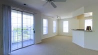 This single-floor space offers a clean, open layout designed to maximize comfort and natural light. The main living area is finished with neutral-toned wall-to-wall carpeting that adds warmth underfoot, while soft cream-colored walls create a calm and inviting atmosphere throughout.

Large windows on multiple walls flood the interior with abundant natural light, enhancing the sense of openness. A glass sliding door with a decorative iron security gate provides direct access to a private outdoor balcony, where a white railing frames views of the surrounding neighborhood — an ideal spot for enjoying fresh air.

Overhead, a ceiling fan with integrated light fixture provides both airflow and illumination, while a separate track lighting fixture adds targeted accent lighting to another area of the room. A partial countertop or breakfast bar is visible along one wall, suggesting a kitchen or kitchenette area that flows seamlessly into the main living space.

The overall layout is open and versatile, with no interior walls dividing the main area, making it well-suited for a variety of furniture arrangements. The space combines practical features with a light, airy aesthetic, resulting in a comfortable and functional single-floor living environment.