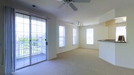 This single-floor space offers a clean, open layout designed to maximize comfort and natural light. The main living area is finished with neutral-toned wall-to-wall carpeting that adds warmth underfoot, while soft cream-colored walls create a calm and inviting atmosphere throughout.

Large windows on multiple walls flood the interior with abundant natural light, enhancing the sense of openness. A glass sliding door with a decorative iron security gate provides direct access to a private outdoor balcony, where a white railing frames views of the surrounding neighborhood — an ideal spot for enjoying fresh air.

Overhead, a ceiling fan with integrated light fixture provides both airflow and illumination, while a separate track lighting fixture adds targeted accent lighting to another area of the room. A partial countertop or breakfast bar is visible along one wall, suggesting a kitchen or kitchenette area that flows seamlessly into the main living space.

The overall layout is open and versatile, with no interior walls dividing the main area, making it well-suited for a variety of furniture arrangements. The space combines practical features with a light, airy aesthetic, resulting in a comfortable and functional single-floor living environment.