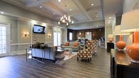 This thoughtfully designed single-floor communal space exudes a sophisticated yet welcoming ambiance, blending upscale finishes with comfortable furnishings. The centerpiece of the room is a striking coffered ceiling adorned with a multi-light chandelier that casts warm, inviting illumination throughout the open layout. Rich hardwood-style flooring runs the length of the space, anchoring the interior with a sense of warmth and continuity.

The main lounge area features a well-curated mix of seating, including a neutral sectional sofa accented with vibrant orange throw pillows and a pair of bold, patterned accent chairs in orange and white geometric print. A low-profile coffee table and decorative area rug define the seating arrangement, creating a cohesive social hub.

One wall is highlighted by a stacked stone accent panel framing a wall-mounted flat-screen television, flanked by subtle sconce lighting that adds depth and texture to the design. French doors along the same wall invite natural light into the interior. Toward the rear of the space, a darker stone-clad wall frames what appears to be a service or refreshment counter with pendant lighting above, suggesting a café or concierge-style amenity area. The overall atmosphere is polished, modern, and well-appointed.