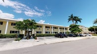 This single-floor building presents a clean and inviting Florida-style aesthetic, characterized by a warm yellow stucco exterior accented with crisp white columns that line the front facade. The architectural design reflects a classic, colonial-inspired style that is both timeless and well-suited to its tropical surroundings. The exterior is framed by mature trees, manicured hedgerows, and swaying palm trees, contributing to a lush, verdant atmosphere that is quintessentially South Floridian. Large windows with dark shutters allow for ample natural light throughout the interior while maintaining a cohesive and polished exterior appearance. A covered colonnade runs along the front of the building, providing shade and a welcoming sense of arrival. Convenient on-site parking is available directly in front of the building, ensuring easy accessibility for occupants and visitors alike. The surrounding landscape, filled with tropical greenery set against vivid blue skies, enhances the overall ambiance of the property, creating a serene and pleasant environment. The building's well-maintained condition and thoughtful design details make it a standout example of classic Florida commercial or multi-use architecture.