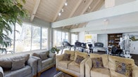 Located at 5550 Genesee Avenue in San Diego, California, this single-story community space serves as both a social lounge and fitness center. The interior is immediately defined by its striking vaulted wood-plank ceiling, supported by exposed beams and fitted with track lighting that illuminates the entire area with a warm, inviting glow. Large windows line the walls, flooding the space with generous natural light and offering views of the surrounding landscape and outdoor greenery.

The lounge area is furnished with comfortable sofas and an armchair arranged on a area rug, creating a welcoming and relaxed gathering space for residents. Adjacent to the lounge, a well-equipped fitness zone features cardio machines including treadmills and ellipticals, providing a convenient workout area within the same open-plan layout. The seamless flow between the lounge and fitness areas makes the space feel expansive and versatile.

The overall ambiance is light, airy, and casual, with the natural wood ceiling and large windows contributing to a bright, open atmosphere. The combination of social seating, exercise equipment, and architectural character makes this a multifunctional community hub designed to accommodate both relaxation and active lifestyles.