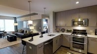 This single-floor residence showcases a thoughtfully designed open-concept layout that integrates the kitchen, dining, and living areas into one cohesive space. The kitchen serves as the focal point, featuring flat-front cabinetry in a warm taupe-gray finish with brushed nickel hardware, complemented by crisp white countertops and a stainless steel undermount sink. Stainless steel appliances, including a double oven range, microwave, and dishwasher, add a polished, contemporary feel. A peninsula with bar-height seating and pendant lighting overhead creates an ideal casual dining or entertaining area. The two pendant lights provide warm, focused illumination that enhances the modern aesthetic. The open layout allows a clear sightline into the living area, where dark upholstered seating and subtle decor contribute to a sophisticated ambiance. A glimpse into the bedroom reveals a well-lit, neatly appointed sleeping area. Hardwood-style flooring runs throughout, tying the spaces together cohesively. The overall design balances functionality with modern style, making efficient use of the available space.