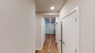 This single-floor space showcases a clean, contemporary design with warm hardwood-style flooring running seamlessly throughout every room, complemented by crisp white walls and recessed lighting that create a bright and welcoming atmosphere.

The kitchen is well-appointed with medium-toned wood cabinetry offering ample storage, white quartz countertops, a classic subway tile backsplash, and a full suite of stainless steel appliances including an electric range, over-the-range microwave, refrigerator, and dishwasher. A stainless steel sink with a modern faucet rounds out the functional workspace.

Adjacent to the kitchen, the open living area provides a generous, versatile space bathed in natural light from a bank of large windows dressed with horizontal blinds. The room offers flexibility for a variety of layout configurations.

A well-lit hallway with recessed lighting and a frosted glass door connects the main living spaces to the private areas of the unit, contributing to the space's sense of flow and openness.

The bathroom is finished with a wood-toned vanity, white quartz countertop, an undermount sink, a large mirror with overhead lighting, a standard toilet, and a shower-tub combination. A towel bar and grab bar add both style and practicality to the space.
