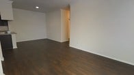 This single-floor space offers a clean, minimalist aesthetic defined by a thoughtful combination of materials and neutral tones. The main living area is anchored by rich, dark hardwood flooring that runs throughout, providing a warm contrast against the crisp white walls and white baseboards. Crown molding trims the upper edges of the walls, adding a subtle architectural detail that elevates the overall finish of the space. Recessed lighting is installed in the ceiling, offering a streamlined lighting solution that keeps the visual lines uncluttered. The open layout of the main area allows for flexible furniture arrangement and contributes to a sense of spaciousness. A doorway opening visible along one wall leads to an adjacent area, suggesting a functional flow between rooms. Electrical outlets are positioned along the walls at standard intervals, reflecting practical, move-in-ready construction. The walls are rendered in a light, neutral tone throughout, creating a bright and versatile backdrop suitable for a variety of interior styles. Overall, the space presents a well-maintained, modern interior with durable finishes designed for everyday living.