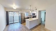 This single-floor residence showcases a thoughtfully designed open-concept layout centered around a contemporary kitchen. The kitchen is a standout feature, finished with a striking two-tone cabinet scheme pairing crisp white upper cabinets with dark lower cabinetry, complemented by quartz countertops and a subtle tile backsplash in a soft gray linear pattern. A full suite of stainless steel appliances—including a refrigerator, range, and over-the-range microwave—provides both style and functionality. Pendant lighting hangs elegantly above the extended kitchen island, which offers additional prep space and a natural gathering point. Track lighting runs overhead, ensuring the workspace is well-lit throughout. Light-toned hardwood-style flooring flows seamlessly throughout the space, lending warmth and continuity to the open layout. A sliding glass door connects the interior to an outdoor balcony, inviting natural light deep into the living area. Additional storage is thoughtfully integrated with built-in cabinetry visible beyond the kitchen. The overall aesthetic is clean, modern, and move-in ready, with every element contributing to a cohesive and functional living environment.