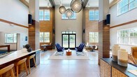 Located at 6 Powder Mill Rd in Maynard, MA, this single-floor amenity space offers a thoughtfully designed layout that seamlessly blends social, recreational, and lounge areas. Soaring vaulted ceilings with warm wood beams define the dramatic living room, which features a stone-clad fireplace with stacked log storage, a mounted flat-screen TV, and plush seating arranged on a patterned area rug. Decorative wall art and blue geometric ceiling paneling add a striking visual character to the space. The open kitchen is outfitted with dark wood cabinetry, white quartz countertops, a herringbone tile backsplash, and a large island with bar seating — ideal for gatherings. The dining area showcases an exposed brick accent wall that rises to full ceiling height, with built-in bench seating and natural light pouring in through large windows overlooking lush greenery. A dedicated games room features a full-size pool table beneath a modern gold pendant light and sliding barn-style doors, while an adjacent room houses a shuffleboard table with a wall-mounted TV. Throughout the space, speckled blue carpet, large-format tile flooring, recessed lighting, and curated furnishings create a cohesive, upscale atmosphere.