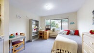 Located on the fifth floor at 210 Dryden Rd in Ithaca, NY, this studio apartment offers a practical and well-organized single-floor layout designed for comfortable everyday living. The entry opens directly into a galley-style kitchen, which is equipped with a full-size refrigerator, electric range, microwave, and ample cream-colored cabinetry providing generous storage. A compact dining table with seating sits adjacent to the kitchen, creating a convenient eat-in area. The main living and sleeping space is carpeted and bathed in natural light through a large window fitted with an air conditioning unit, offering views of the surrounding Ithaca neighborhood and treetops. This versatile room accommodates a full-size bed, a wooden study desk with a filing drawer unit, and a freestanding bookshelf, making it well-suited for both rest and focused work. Built-in shelving along one wall adds additional storage and display space. A private bathroom is accessible directly from the main room, featuring a vanity with sink and mirror. The unit's light-toned walls and neutral carpet contribute to a clean, functional atmosphere throughout. This apartment is ideally situated near Cornell University and Ithaca College, making it a practical choice for students or young professionals.