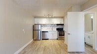 This single-floor unit offers a clean, modern interior with consistent light wood-style luxury vinyl flooring flowing seamlessly throughout every room. The kitchen is a standout feature, showcasing white shaker-style cabinetry with brushed nickel hardware, a stylish mosaic tile backsplash in neutral tones, and a full suite of stainless steel appliances including a refrigerator, dishwasher, and electric range. A deep stainless steel sink with a contemporary faucet and light-toned countertops complete the kitchen's polished aesthetic. Track lighting overhead adds focused illumination to the cooking space. The open layout allows the living area to extend naturally from the kitchen, creating a functional and connected floor plan. The main living space is bright and airy, featuring a ceiling fan with a light fixture and a wall-mounted HVAC unit for year-round climate control. A window allows natural light to filter in, enhancing the room's open feel. The bathroom is efficiently designed with white tilework, a full-sized tub and shower combination, a toilet, and a white vanity with ample counter space. Light-toned flooring continues into the bathroom, maintaining a cohesive look throughout. The overall aesthetic is fresh, neutral, and move-in ready.