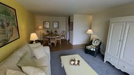 This single-floor residence offers an open and functional layout, with the living and dining areas flowing seamlessly into one another. The living area features plush carpeting underfoot, a comfortable sofa, and a wicker accent chair with tropical-print upholstery, contributing a relaxed, casual atmosphere. A white-painted coffee table serves as a central focal point, styled with simple decorative accents. Warm lighting from floor and table lamps adds a cozy ambiance throughout the space. The dining area transitions naturally from the living room and is defined by rich hardwood-style flooring. A round dining table surrounded by rattan-style chairs provides an intimate setting for meals, complemented by framed artwork on the neutral walls and a classic floor lamp. A partial view into an adjacent area — likely a kitchen or hallway — suggests additional functional spaces beyond what is immediately visible. The overall palette of warm beiges and soft neutrals, combined with the mix of carpet and hardwood flooring, creates a cohesive and inviting interior throughout this well-laid-out single-level home.