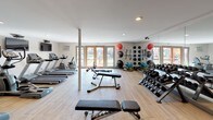Located at 9128 Burke St in Pico Rivera, California, this single-floor fitness center is a well-appointed exercise facility designed to support a comprehensive workout experience. The space features light wood-style flooring throughout, lending a clean and modern aesthetic that complements the neutral white walls and recessed lighting overhead.

The cardio zone is anchored by a row of treadmills and elliptical trainers, each equipped with individual flat-screen televisions mounted on the wall for an engaging workout experience. Adjacent to the cardio equipment, a strength training area houses a full dumbbell rack with a wide range of weights, multiple adjustable weight benches, and a multi-station cable machine, providing options for a full-body resistance workout.

Floor-to-ceiling mirrored walls on one side of the room visually expand the space while allowing users to monitor their form during exercise. A dedicated functional fitness area includes medicine balls, stability balls, resistance bands, and additional accessories neatly organized on wall-mounted racks.

Natural light filters in through large glass French doors that open to an outdoor area, creating a bright and inviting atmosphere. The facility also includes wall-mounted televisions, a support column in the center of the space, and a security camera, reflecting a thoughtfully designed environment built for both functionality and comfort.