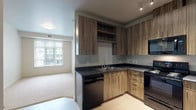 This single-floor residential unit offers a thoughtfully designed layout with modern finishes and a cohesive neutral color palette. The kitchen is a standout feature, showcasing wood-grain cabinetry in a warm walnut tone paired with dark countertops and light vinyl plank flooring. A full suite of black appliances — including a range, over-the-range microwave, dishwasher, and refrigerator — provides complete functionality. Upper and lower cabinetry offer generous storage, and an open shelf adds a contemporary touch. The kitchen opens directly to the living area, creating a seamless flow between spaces. The living and bedroom areas are finished with plush wall-to-wall carpeting in a soft neutral tone, contributing to a warm and comfortable ambiance. Multiple windows fitted with vertical blinds allow natural light to fill these rooms while maintaining privacy. The bedroom spaces are generously proportioned, each offering ample room and natural light. The bathroom features light vinyl plank flooring consistent with the kitchen, a wide vanity with dark countertop and wood-grain cabinetry, a large mirror, and a bathtub. Chrome fixtures and a well-lit vanity complete the clean, functional design. Overall, the unit presents a modern, move-in-ready aesthetic with practical, quality finishes throughout.