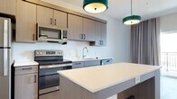 This thoughtfully designed single-floor unit showcases a cohesive modern aesthetic with light wood-look flooring and a neutral color palette carried throughout the space. The open-concept kitchen serves as a central highlight, featuring flat-front cabinetry in a warm taupe wood finish with matte black hardware, crisp white quartz countertops, and a full suite of stainless steel appliances including a refrigerator, electric range, over-the-range microwave, and dishwasher. A generously sized kitchen island with built-in shelving provides additional prep space and storage, while a pair of distinctive teal pendant lights add a bold pop of color and contemporary character overhead. The bathroom is finished to a high standard, offering a wood-grain vanity with open shelving, white quartz countertop, an undermount sink with a modern chrome faucet, and an illuminated mirror cabinet above. Light wood-look tile flooring completes the clean, cohesive look. The bedroom is a calm and understated retreat, featuring the same warm wood-look flooring, crisp white walls, and a window dressed with neutral drapes and blinds, allowing for flexible light control. The overall ambiance of the unit is clean, contemporary, and move-in ready.