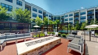 Located at 548 Lancaster Ave in Malvern, PA, this meticulously designed outdoor courtyard amenity space is set within a modern multi-story residential building featuring a contemporary façade of neutral siding, stone accents, and large dark-framed windows. The centerpiece of the courtyard is a generously sized resort-style swimming pool with an adjoining spa, surrounded by a wide pool deck lined with lounge chairs and umbrella-shaded seating areas, ideal for relaxing under open skies. Adjacent to the pool, pergola structures with slatted wood screens define intimate seating zones, while a prominent stone fire pit with a river rock feature serves as a social focal point, surrounded by sectional seating and accent chairs. A dedicated grilling and dining area includes multiple built-in gas grills set against a brick backdrop, accompanied by outdoor dining tables and fire pit seating for al fresco gatherings. A bocce ball court and putting green offer recreational variety. Warm-toned brick pavers and natural wood decking connect the various zones, complemented by abundant, mature landscaping including ornamental grasses, evergreen shrubs, and deciduous trees that provide shade and greenery throughout. String lights overhead add a welcoming ambiance to the entire courtyard, creating a well-appointed outdoor environment designed for both activity and relaxation.