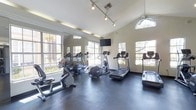 This fitness center offers a clean, well-organized space designed to meet a variety of cardio and exercise needs. The room features vaulted ceilings that lend an open, airy feel, complemented by an abundance of natural light streaming in through large, white-framed windows along multiple walls. A ceiling fan aids in ventilation and comfort during workouts. The flooring consists of durable rubber mat tiles, providing traction and impact absorption for exercisers. A full-length wall mirror on one side visually expands the space and allows users to monitor their form. The equipment lineup includes multiple treadmills, an elliptical trainer, a stationary upright bike, and an additional cardio machine, offering a well-rounded selection for cardiovascular training. Track lighting runs along the ceiling, providing even, directed illumination throughout the room. A wall-mounted flat-screen television is positioned for visibility from the cardio machines, adding an entertainment option during workouts. The overall ambiance is bright, functional, and inviting, reflecting a thoughtfully designed facility intended to encourage regular fitness activity.