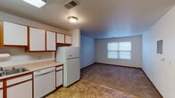 This single-floor unit offers a practical and well-organized layout designed for comfortable everyday living. The kitchen is a standout functional space, featuring white cabinetry with warm wood trim accents, light-toned countertops, and tile flooring. It is fully equipped with a white electric range, dishwasher, and refrigerator, along with ample upper and lower cabinet storage and a double-basin sink positioned beneath additional cabinetry. The kitchen flows naturally into the living area, which features plush carpeting, neutral white walls, and a window that allows natural light to fill the room. A thermostat and wall vents indicate a central climate control system throughout the unit. The bedroom is a quiet, carpeted room with a window fitted with blinds, providing both privacy and natural light. The bathroom is notably designed with accessibility in mind, featuring stainless steel grab bars near the toilet and shower area, a vanity with generous counter space and storage drawers, a large mirror with overhead lighting, and durable tile flooring. Throughout the unit, warm wood-toned door frames and baseboards provide a cohesive design element, tying the spaces together into a clean, functional, and accessible living environment.