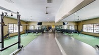 This fitness center in Flagstaff, Arizona offers a generously sized, single-floor exercise space designed to accommodate a wide range of workout styles and fitness goals. The open layout is divided into distinct functional zones, each thoughtfully outfitted with professional-grade equipment.

The cardio area features a row of treadmills positioned in front of large windows, allowing natural light to fill the space and providing views of the surrounding exterior. A large flat-screen TV is mounted on the wall nearby for entertainment during workouts. A heavy-duty tire and additional cardio machines, including stationary bikes and a stair climber, round out this section.

The strength and free weights area includes multiple dumbbell racks with a full range of weights, barbells, a multi-functional power rack, and adjustable benches. Full-length mirrors line the walls, enhancing the sense of space and allowing users to monitor their form. Artificial turf flooring designates a dedicated functional training zone, ideal for sled work, agility drills, and floor exercises.

Ceiling fans provide air circulation throughout, complementing the recessed lighting that ensures the space is bright and well-lit. The durable rubber and turf flooring combination offers both safety and versatility. The warm, neutral-toned walls and clean design create a motivating and professional training environment.