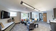 This single-floor facility is thoughtfully designed to offer a range of amenity spaces within a cohesive, modern aesthetic. The expansive lounge area serves as the centerpiece of the space, furnished with multiple seating arrangements featuring gray upholstered sofas and accent chairs accented with colorful throw pillows. Large windows allow generous natural light to flood the interior, offering views of mature trees outside. Overhead, industrial-style linear lighting fixtures complement the neutral gray flooring, which runs consistently throughout the space. A wall-mounted flat-screen television and built-in ledge provide additional functionality to the lounge area.

Adjacent to the lounge, a dining area with black chairs and a table offers a secondary gathering space, while colorful artwork adds visual interest to the walls. The kitchenette features gray cabinetry, a white countertop, stainless steel sink, a beverage refrigerator, and a striking reclaimed wood accent wall that introduces warmth and texture to the space.

The fitness center is well-appointed with cardio machines, a cable weight system, a dumbbell rack, and a weight bench. Exposed ductwork on the high ceilings adds an industrial character, while a large, vibrant mural and floor-to-ceiling mirrors energize the workout environment. Glass double doors provide direct outdoor access.