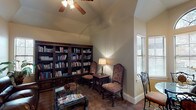 This library is a thoughtfully designed space that balances functionality with refined, traditional aesthetics. The room is anchored by a dramatic vaulted ceiling, which lends an airy sense of height and openness, complemented by a ceiling fan that adds comfort and casual charm. The flooring features a distinctive diamond-patterned tile that adds visual interest throughout.

A floor-to-ceiling built-in bookcase constructed of dark, ornately carved wood dominates one wall, offering generous shelving capacity and serving as both a functional and decorative focal point. The adjoining reading area includes upholstered high-back chairs and a side table with a classic table lamp, creating an intimate setting ideal for quiet reading.

A rich dark leather sofa anchors the seating area, paired with a glass-top coffee table. Framed artwork adds a decorative accent on the neutral-toned walls. A standout feature is the bay window nook bathed in natural light, where an arched transom window crowns a set of large windows with blinds. A round glass-top table with ornate wrought-iron chairs occupies this bright corner.

The entry corridor leading to the library features elegant white columns with classical detailing, tile flooring, and an adjacent dining area, establishing a grand sense of arrival.