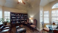This library is a thoughtfully designed space that balances functionality with refined, traditional aesthetics. The room is anchored by a dramatic vaulted ceiling, which lends an airy sense of height and openness, complemented by a ceiling fan that adds comfort and casual charm. The flooring features a distinctive diamond-patterned tile that adds visual interest throughout.

A floor-to-ceiling built-in bookcase constructed of dark, ornately carved wood dominates one wall, offering generous shelving capacity and serving as both a functional and decorative focal point. The adjoining reading area includes upholstered high-back chairs and a side table with a classic table lamp, creating an intimate setting ideal for quiet reading.

A rich dark leather sofa anchors the seating area, paired with a glass-top coffee table. Framed artwork adds a decorative accent on the neutral-toned walls. A standout feature is the bay window nook bathed in natural light, where an arched transom window crowns a set of large windows with blinds. A round glass-top table with ornate wrought-iron chairs occupies this bright corner.

The entry corridor leading to the library features elegant white columns with classical detailing, tile flooring, and an adjacent dining area, establishing a grand sense of arrival.