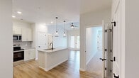 This well-designed 3-bedroom, 2-bathroom single-floor home at 1215 Evolve Way in Greenville, NC offers a modern, cohesive layout that maximizes both functionality and comfort. The open-concept living area flows seamlessly from the kitchen, which serves as the heart of the home. The kitchen is outfitted with white shaker cabinetry, a decorative tile backsplash, granite countertops, stainless steel appliances — including a refrigerator with ice maker, range, microwave, and dishwasher — and a central island with a matte black sink and stylish pendant lighting overhead. Light hardwood-style flooring extends throughout the main living areas, complemented by a ceiling fan and natural light from balcony-facing glass doors. The three bedrooms feature plush carpeting, ceiling fans, and windows with outdoor views. The spacious primary bedroom includes its own closet. Both bathrooms are finished with granite-topped vanities, undermount sinks, and matte black fixtures; one features a glass-enclosed shower, the other a full tub-shower combo. A dedicated laundry room houses full-size washer and dryer units with overhead shelving. Three closets and a hallway complete this thoughtfully arranged home.