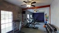 This single-floor fitness center offers a dedicated space for cardio and general exercise, thoughtfully designed to accommodate a range of workout routines. The room features a collection of cardio equipment, including a treadmill, elliptical trainers, and a stationary bike, all arranged to allow comfortable use and movement throughout the space. The flooring is carpeted, with protective rubber mats placed beneath the equipment to preserve the surface and provide stability during workouts. Two ceiling fans are mounted overhead, ensuring consistent airflow and comfort during exercise sessions. The walls are painted in a combination of gray and deep navy blue, with bold red accent trim adding a dynamic, energetic feel to the environment. A wall-mounted television is positioned for easy viewing, making workouts more engaging. Natural light filters in through a window, brightening the space during daytime hours. Framed artwork and motivational posters decorate the walls, contributing to an encouraging atmosphere. Exercise accessories, including balance balls and free weights, are also present, rounding out the available fitness options. Overall, this fitness center provides a functional and well-appointed environment suited for consistent, varied physical activity.
