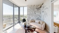 Located at 9600 S Dixie Hwy in Miami's Pinecrest corridor, this high-rise residence offers a refined contemporary living experience defined by clean lines, a neutral palette, and sweeping panoramic views of the Miami skyline and lush greenery below. Floor-to-ceiling glass windows and sliding doors flood the interior with natural light and open onto a private balcony, creating a seamless indoor-outdoor connection throughout the living area.

The living space features a striking tropical-themed mural wallcovering alongside expansive corner glazing, lending the room both artistic character and a strong sense of openness. The kitchen is outfitted with flat-front wood-grain cabinetry, stainless steel appliances including a full-size refrigerator, range, microwave, and dishwasher, and crisp white countertops with an island for additional workspace.

Each bedroom is thoughtfully appointed with custom accent wallcoverings — one showcasing a bold botanical monstera leaf mural and another featuring an organic abstract pattern — paired with large windows that frame verdant city views. Light wood-tone flooring runs throughout, adding warmth to the contemporary aesthetic.

The bathrooms are finished with neutral tile, glass-enclosed walk-in showers with rain showerheads, and clean white vanities, maintaining a consistent sense of modern elegance throughout the residence.