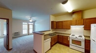 This single-floor unit is thoughtfully designed with an open-concept layout that connects the kitchen and living areas seamlessly. The kitchen is well-equipped with warm honey-toned wood cabinetry, dark laminate countertops, a white electric range, dishwasher, and refrigerator. A peninsula with a breakfast bar separates the kitchen from the living space, enhancing functionality. Tile flooring runs throughout the kitchen, while plush carpet extends across the living and bedroom areas.

The living room is generously proportioned, featuring a ceiling fan and access to a private balcony through a glass door, allowing natural light to fill the space. Multiple bedrooms are present, each carpeted and outfitted with ceiling fans and baseboard heating. Windows in each bedroom provide natural light and outdoor views, contributing to a bright and comfortable atmosphere.

The bathroom is designed with accessibility in mind, featuring grab bars near the toilet, a wall-mounted vanity with warm wood cabinetry, a lighted mirror, and tile flooring. The overall aesthetic throughout the unit is neutral and clean, with warm wood accents complementing the light-toned walls, creating a welcoming and practical living environment.