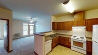 This single-floor unit is thoughtfully designed with an open-concept layout that connects the kitchen and living areas seamlessly. The kitchen is well-equipped with warm honey-toned wood cabinetry, dark laminate countertops, a white electric range, dishwasher, and refrigerator. A peninsula with a breakfast bar separates the kitchen from the living space, enhancing functionality. Tile flooring runs throughout the kitchen, while plush carpet extends across the living and bedroom areas.

The living room is generously proportioned, featuring a ceiling fan and access to a private balcony through a glass door, allowing natural light to fill the space. Multiple bedrooms are present, each carpeted and outfitted with ceiling fans and baseboard heating. Windows in each bedroom provide natural light and outdoor views, contributing to a bright and comfortable atmosphere.

The bathroom is designed with accessibility in mind, featuring grab bars near the toilet, a wall-mounted vanity with warm wood cabinetry, a lighted mirror, and tile flooring. The overall aesthetic throughout the unit is neutral and clean, with warm wood accents complementing the light-toned walls, creating a welcoming and practical living environment.