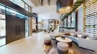 Located at 2101 Bond Street in Charlottesville, Virginia, this striking lobby space delivers a refined, contemporary atmosphere across a single, expansive floor. The design is defined by soaring double-height ceilings adorned with bold exposed beams and dramatic pendant lighting fixtures, including large drum-shaped pendants with warm metallic interiors that cast an inviting glow throughout the space. Floor-to-ceiling curtain-wall windows line the perimeter, flooding the interior with abundant natural light and offering clear views of the surrounding streetscape. The flooring transitions seamlessly between large-format light stone tile and warm wood plank sections, subtly delineating distinct seating zones. Multiple thoughtfully arranged lounge areas are furnished with contemporary sofas, curved sectionals, and accent chairs in a neutral palette of blues, grays, and warm earth tones, anchored by layered area rugs. Textural stone accent walls, slatted wood screens, and backlit decorative wall panels provide rich visual depth and serve as architectural focal points. Integrated greenery in tall planters adds a natural element throughout. A bar or service counter area with bar seating is visible toward the rear, completing this versatile, hospitality-oriented gathering space.