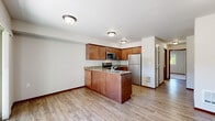 Unit 104 is a well-designed one-bedroom, one-bathroom residence that makes efficient use of its single-floor layout. The open-concept kitchen and living area serves as the heart of the unit, where warm-toned wood cabinetry with a tile backsplash pairs with stainless steel appliances — including a refrigerator, range, microwave, and dishwasher — and a granite-style countertop peninsula with an integrated double sink. Light wood-style vinyl flooring runs throughout the main living areas, creating a cohesive and easy-to-maintain surface. The living room benefits from natural light through windows that offer views of the surrounding exterior. The bedroom is a comfortable, well-proportioned space featuring plush carpet, a large window that brings in ample natural light and an outdoor view, and a sliding-door closet offering generous storage. The bathroom is appointed with warm tile flooring, a wood vanity with granite-style countertop, a large mirror, and a full bathtub with safety bar. The unit's neutral color palette and consistent finishes throughout create a clean, move-in-ready atmosphere suited for a variety of living styles.