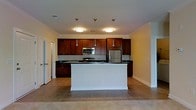 This single-floor space features an open-concept kitchen that serves as the focal point of the layout. The kitchen is outfitted with rich, dark-stained wood cabinetry that extends along the full back wall, providing generous storage capacity. A stainless steel refrigerator and microwave are seamlessly integrated alongside a range, offering a cohesive and functional appliance suite. Dark countertops contrast elegantly with the lighter cabinetry trim and walls, adding visual depth to the space.

A central white island anchors the kitchen, providing additional prep space and serving as a natural dividing element between the kitchen and the surrounding open area. Pendant lighting hangs above the island, complementing the flush-mount ceiling fixtures that illuminate the space evenly. The flooring throughout consists of neutral-toned ceramic tiles, lending a clean and durable foundation to the area.

Crown molding runs along the ceiling perimeter, adding an architectural detail that elevates the overall finish of the space. The walls are painted in a soft, warm neutral tone, contributing to a bright and welcoming ambiance. A doorway visible to the left suggests additional rooms beyond the main open area.