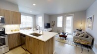 This single-floor unit offers a thoughtfully designed open-concept layout that connects the kitchen and living areas into one cohesive, functional space. The kitchen serves as the centerpiece, anchored by a large island with a white quartz countertop and an undermount stainless steel sink, complemented by warm wood-toned cabinetry and stainless steel appliances, including a dishwasher. Recessed lighting overhead ensures the kitchen remains well-lit at all times. Light wood-style flooring flows continuously throughout the space, contributing to a clean, contemporary aesthetic. The living area is generously proportioned and bathed in natural light, thanks to multiple large windows dressed with white horizontal blinds. A glass-paneled exterior door provides additional light and access to the outdoors. The neutral gray wall palette creates a calm, versatile backdrop that complements the modern finishes. A wall-mounted media console and a comfortable seating arrangement make the living space both practical and inviting. The overall ambiance is bright, airy, and modern, with an emphasis on open sightlines and a seamless flow between everyday living spaces.