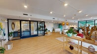 This bright and thoughtfully designed single-floor space seamlessly blends contemporary style with a relaxed, coastal-inspired aesthetic. The open floor plan is anchored by warm-toned hardwood-style flooring that flows throughout, connecting each functional zone with visual continuity.

The main lounge area is a standout feature, furnished with a generous white sectional sofa, vibrant coral ottomans, and a dark coffee table set atop a textured area rug. A wall-mounted flat-screen television, open shelving with curated decor, and an assortment of lush tropical plants contribute to the inviting ambiance. Large black-framed floor-to-ceiling glass doors and windows flood this area with natural light and provide views of an outdoor terrace.

Adjacent to the lounge, a stylish kitchenette commands attention with its eye-catching mosaic tile backsplash in soft blue-grey tones, sleek white cabinetry, light stone countertops, and a retro-style turquoise refrigerator that serves as a bold focal point. A terrazzo-finished curved counter adds an artistic touch to the space.

The entry zone features dramatic navy-framed glass double doors, creating a striking architectural statement. Track lighting runs throughout the ceiling, providing flexible and well-distributed illumination. Abundant greenery and carefully selected decorative accents tie the space together, resulting in a warm, welcoming, and visually dynamic environment.