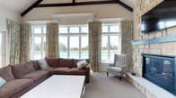 Located at 3381 Blue Heron Dr in Hilliard, Ohio, this single-floor community gathering space combines refined architectural details with a warm, inviting atmosphere. The room is anchored by soaring vaulted ceilings accented with rich, dark exposed wood beams that add a rustic yet elegant character to the space. Large, floor-to-ceiling windows line the wall, flooding the interior with an abundance of natural light and offering unobstructed views of the outdoor pool area beyond — creating a seamless connection between indoor comfort and outdoor recreation. Dressed with floor-length floral drapery panels, the windows add a soft, classic touch to the overall design. A stone fireplace anchors one side of the room, providing a cozy focal point that balances the openness of the windowed wall. The lounge area is furnished with a generous sectional sofa in warm brown leather complemented by a neutral upholstered armchair, arranged around a clean-lined white coffee table — ideal for relaxed gatherings or casual socializing. Neutral carpeting and cream-toned walls maintain a light, airy feel throughout. The combination of architectural craftsmanship, natural light, and thoughtful furnishings makes this a comfortable and functional space for community members to gather and unwind.