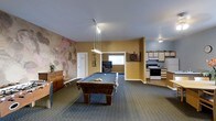 This single-floor space is designed with entertainment and functionality in mind. The main area is anchored by a full-sized pool table positioned beneath a pendant light fixture that provides focused overhead illumination. The room features carpeted flooring and warm yellow-toned walls, creating an inviting atmosphere. A textured accent wall with an abstract, earthy mural adds a distinctive artistic element to the space.

A dartboard is mounted on the wall nearby, complementing the recreational theme of the room. Adjacent to the main area, a lounge nook houses a television and seating, offering a comfortable spot for relaxing between games. A window in the lounge area allows natural light to filter in.

To one side, a kitchenette provides practical amenities including a white range, upper cabinetry, and counter space — making the space well-suited for hosting gatherings. A wooden dresser and decorative plant add warmth and character to the overall design. The layout is open and spacious, allowing for easy movement between the recreational, lounge, and kitchen zones.