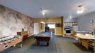 This single-floor space is designed with entertainment and functionality in mind. The main area is anchored by a full-sized pool table positioned beneath a pendant light fixture that provides focused overhead illumination. The room features carpeted flooring and warm yellow-toned walls, creating an inviting atmosphere. A textured accent wall with an abstract, earthy mural adds a distinctive artistic element to the space.

A dartboard is mounted on the wall nearby, complementing the recreational theme of the room. Adjacent to the main area, a lounge nook houses a television and seating, offering a comfortable spot for relaxing between games. A window in the lounge area allows natural light to filter in.

To one side, a kitchenette provides practical amenities including a white range, upper cabinetry, and counter space — making the space well-suited for hosting gatherings. A wooden dresser and decorative plant add warmth and character to the overall design. The layout is open and spacious, allowing for easy movement between the recreational, lounge, and kitchen zones.