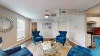 This single-floor space is thoughtfully designed to balance comfort and functionality, offering a warm and inviting atmosphere throughout. The main lounge area serves as the centerpiece, featuring rich teal velvet wingback chairs arranged around a round glass-and-gold coffee table atop a neutral area rug. Soft grey walls, recessed lighting, and a ceiling fan contribute to the room's polished yet relaxed ambiance. Dark hardwood-style flooring runs consistently throughout, unifying the space visually.

Natural light flows in generously through windows fitted with clean white blinds, and a set of glass-paned French doors at the entry provides an open, accessible feel while offering views of the surrounding outdoor greenery. A decorative sunburst mirror accents the entryway wall, adding an elegant touch.

The kitchen is well-appointed with white shaker-style cabinetry, light granite countertops, a stainless steel farmhouse sink, and a stainless steel range. A grey subway tile backsplash adds texture and visual interest, complementing the crisp cabinetry.

Adjacent to the main lounge, a dedicated dining nook features a round wooden table surrounded by coordinating blue velvet chairs with gold-tipped legs. A large abstract art piece in cool blue and white tones anchors the wall, reinforcing the space's cohesive color palette. This space is well-suited for meetings, gatherings, or professional use.