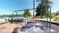 This outdoor playground offers a welcoming recreational space set within a lush, tree-lined environment. The centerpiece of the area is a sturdy green metal swing set accommodating multiple riders, complete with rubber safety matting beneath each swing and a wood chip ground surface to cushion activity. Towering mature trees, including tall pines, provide natural shade throughout the space, creating a comfortable environment on sunny days.

The playground is situated adjacent to a community swimming pool, visible in the background and enclosed by a chain-link fence. Pool lounge chairs and aquatic equipment are visible poolside, indicating a well-equipped aquatic facility just steps away. A small brick utility structure sits nearby on the property grounds, adding a functional element to the overall layout.

The surrounding landscape features a generously sized, well-kept grassy lawn that provides ample open space for additional outdoor activities. Paved walkways wind through the area, offering easy access throughout the grounds. Lush mixed foliage, including magnolia trees and other mature deciduous trees, frames the perimeter and contributes to the green, park-like atmosphere. The entire space benefits from abundant natural light and open sky views, making it an inviting and functional outdoor amenity for community residents.